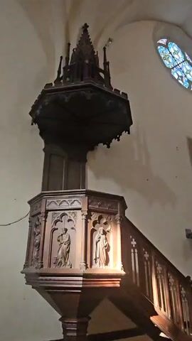 A wooden pulpit with stairs and ornate carvings is shown in a church. The pulpit is dark wood and has a hexagonal base with figures carved into it. The stairs are also made of dark wood and have decorative crosses on the railing. A stained glass window is visible in the background, as well as a painting of Jesus on the cross. The church interior is lit by natural light from the windows and some artificial lighting. The overall impression is one of historical significance and religious solemnity.
