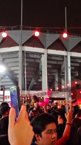 The video captures a vibrant outdoor scene at night, likely during a festival or concert. The foreground shows a crowd of people, many of whom are holding up their smartphones to record the event. Several individuals are visible, including some with raised arms.  Behind the crowd, a stadium stands tall, lit by various red, blue, and green lights strung up on strings.  A large white banner with what appears to be black and white images is also visible.  The atmosphere seems lively and festive, judging by the music and the general excitement of the crowd.