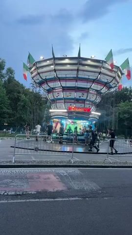 The video shows a carnival ride with people on it. The ride is a spinning carousel with chairs suspended from chains. Italian flags are attached to the top of the ride. The ride is lit up with bright lights, and there are trees in the background. The sky is overcast. Some people are standing around the ride watching. A street is visible in the foreground.