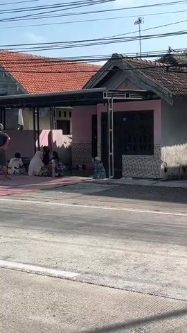 The video shows a street scene with several people gathered outside a pink building. The building has a covered porch area with metal supports. A man in a dark t-shirt and shorts stands facing away from the camera, his hands on his hips. Several other individuals are seated on the ground or on low benches under the porch. Some are wearing casual clothing, while others are wearing headscarves. The ground in front of the building is covered with a red mat. A street runs in front of the building, with a white line painted on it. In the background, there are houses with red tiled roofs and power lines. One house has a sign with yellow lettering and the number 10,000. The sky is clear and blue.