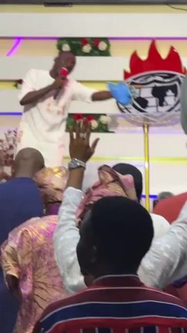 The video shows a man in a white outfit speaking into a microphone while standing behind a clear podium. He is on a stage with decorative elements behind him. In front of the stage, several people are visible from behind, with their hands raised. The atmosphere appears to be a religious service or gathering, indicated by the raised hands and the speaker's presence on a raised platform. The man behind the podium is animated and gesturing, suggesting he is actively engaging with the audience. The lighting is bright, illuminating the stage and the audience.
