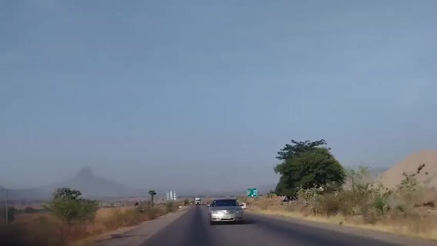 Certainly! Here is a forensic description of the video:

**Scene:** The video depicts a front-view perspective from a moving vehicle on a paved, two-lane road.

**Environment:** The surrounding environment appears dry and arid, with sparse vegetation consisting of shrubs and small trees. In the distance, a hazy mountain peak is visible.

**Objects:**
*   **Vehicles:** A silver sedan is traveling in the opposite direction, towards the camera. A larger, possibly white, vehicle is further down the road in the same direction as the silver sedan.
*   **Utility Pole:** A utility pole is on the left side of the road.

**Actions:**
*   The primary vehicle is moving forward on the road.
*   The silver sedan is moving in the opposite direction.

**Interactions:** There's no direct interaction shown, only the relative movement of the vehicles.

**People:** There are no visible people in the frame.