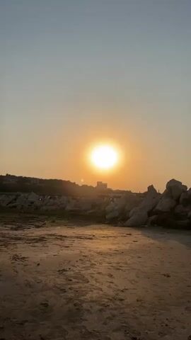 The video shows a sunset over a rocky beach. The sun is a bright orange orb in the center of the frame, partially obscured by a building on the horizon. The sky is a pale orange and blue gradient. The foreground consists of sand and rocks, with a scattering of debris. The tide appears to be out, revealing wet sand.