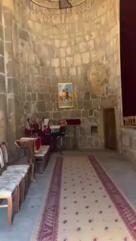 Inside a small church, three women in red robes are performing music. One plays an electronic keyboard, another plays a woodwind instrument, and the third sings. The church has high stone walls in a rounded shape and a decorative chandelier. A red rug runs down the center aisle. A framed picture hangs on the wall behind the performers. Several wooden chairs are lined up on the left side of the aisle.