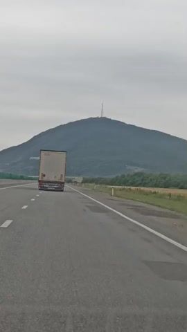 A semi-trailer truck drives on a highway towards a large, tree-covered mountain. The sky is overcast. The truck is white with a red stripe. The highway has a white dashed line on the left and a solid white line on the right. There are fields of dry grass and green bushes on the right side of the road, with more trees and hills in the distance. A cell tower is visible at the top of the mountain.