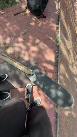The video shows a first-person perspective of someone standing on a skateboard on a brick patio. The skateboard is positioned on a wooden ramp. Two other people are visible in the foreground, also on the patio. One is wearing black pants and black shoes with white soles, standing on a small wooden block. The other is wearing black pants and black shoes with white soles, standing next to the first person.

The scene appears to be outdoors, with dappled sunlight filtering through trees, suggesting it is daytime. The brick patio has some sparse vegetation growing in the cracks. A black backpack is visible in the background. The overall impression is of a casual, possibly recreational setting, where skateboarding might be taking place. There is no visible text in the video.