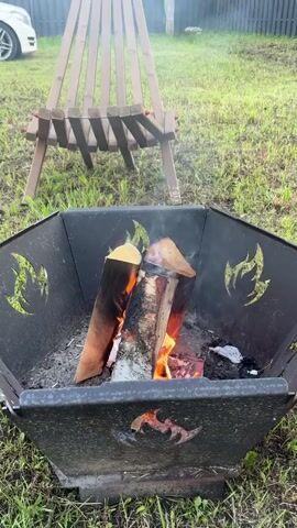A shot of a fire pit with burning logs. The fire pit is black and has flame designs cut into the sides. It sits on green grass. In the background, a wooden log holder stands with a white car parked to the left. The fire pit is the main focus.