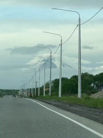 The video shows a long row of streetlights lining a two-lane road. The road is paved and has white lane markers. The streetlights are tall and silver, with curved tops. In the background, a snow-capped mountain is visible, partially obscured by clouds. There are a few cars on the road, and some trees and vegetation on the side. The sky is overcast. The camera appears to be moving forward, possibly from inside a vehicle.