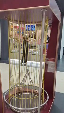 The video opens with a close-up shot of a cylindrical cage-like structure made of yellow cords suspended from a wooden frame. The cage has a metal ring at the bottom and a metal cone-shaped object hanging from the top. In the background, through the cage, a person wearing a dark jacket is walking away from the camera in a shopping mall. The camera then pans to the right, showing more of the shopping mall environment, including store fronts and escalators. One store is labeled "Bijou Brigitte" and displays jewelry and accessories. Another shot shows a sign with German text, possibly an informational display. The video ends with a close-up of a metal ring, similar to the one at the bottom of the cage, resting on a wooden surface. The overall environment appears to be a public indoor space, likely a shopping mall.