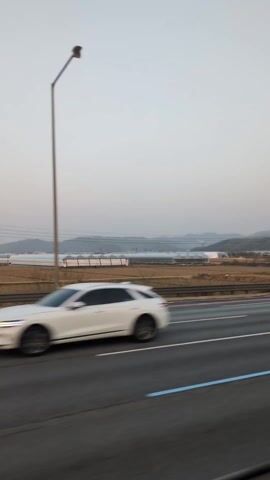 The video shows a view from a moving vehicle on a highway. Various cars pass in the opposite direction, including black sedans, a white SUV, and a white sedan.  The highway has a barrier on the side, and beyond the barrier, there's a field with several greenhouses. Distant mountains are visible under a pale sky.