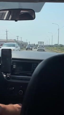 The video shows a view from the inside of a car looking forward. The car is on a road with other vehicles. In the foreground, the dashboard of the car is visible, including a phone mounted on a holder. The road appears to be a two-lane road with traffic moving in the same direction. Several cars and a truck are visible ahead. On the left side of the road, there are buildings and power lines. The sky is clear and the weather appears to be sunny. The overall scene depicts a typical view from a car during a road trip or commute.