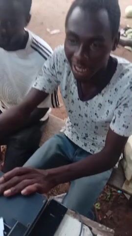 A man is sitting outside, looking at a laptop. He is wearing a white t-shirt with a black pattern and blue jeans. Another man is partially visible to his left. The ground appears to be dirt and there are some green items and a white tarp in the background. The laptop is black with a pink cable connected to it. The man is looking at the laptop and appears to be talking. The second man is out of focus. The scene is lit by daylight.