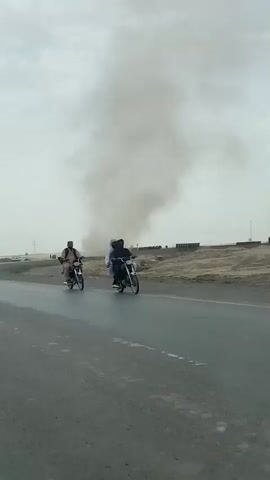 The video captures a large dust devil swirling in the distance, rising high into an overcast sky. The scene is set on a dry, barren landscape, with a two-lane asphalt road in the foreground. On the right side of the frame, a man wearing traditional clothing and a cap is riding a motorcycle away from the camera.

In the middle ground, between the road and the dust devil, there are mounds of earth or sand, possibly eroded. A few structures resembling solar panels are visible on the horizon to the right of the dust devil. Further in the background, to the left of the dust devil, several vehicles, including what appear to be cars and trucks, are seen on another road or track. Utility poles or similar tall structures are also scattered across the distant landscape. The overall atmosphere is hazy and dusty, characteristic of a desert-like environment. No visible text is present in the video.