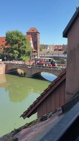A drone shot pans over a bridge adorned with people and a parade float. The bridge spans a green body of water, and on the left bank, lush green trees obscure some of the red-and-white buildings that line the river. The bridge's stone structure is visible, with metal railings decorated with dark figures. Crowds of people are gathered on the bridge, some holding flags and appearing to wave. A large truck-like float, decorated with rainbow colors, balloons, and banners, is proceeding across the bridge. The float has people on it, some of whom are holding flags and interacting with the crowd. The sky is clear and blue. On the right side of the frame, a portion of a building with a brown, shingled roof is visible in the foreground.