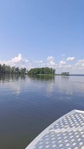 The video opens with a view from a boat looking out over a wide body of calm water. In the distance, there is a small island covered in trees. The sky is clear and blue with a few scattered white clouds. As the video progresses, the boat moves forward, and the island in the distance becomes smaller, while the expanse of water and the horizon become more prominent. A bird can be seen flying in the distance. The camera also captures a glimpse of the boat's edge in the foreground. The scene is bright and appears to be a sunny day on a lake or a similar large body of water.