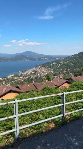 This media file captures a scenic view of a town nestled along a large body of water, with rolling hills and mountains in the background. The scene is bathed in bright sunlight, indicating it is daytime, likely late morning or early afternoon given the strong shadows and vibrant colors. The sky is a clear, brilliant blue with a few scattered white clouds. The weather appears to be warm and pleasant, typical of a summer day in a Mediterranean-like climate.

In the foreground, a metal railing runs horizontally across the lower portion of the frame, partially obscuring a dense growth of green ferns and bushes. Beyond the foliage, several houses with terracotta tile roofs are visible, suggesting a residential area. These houses are clustered on a hillside, overlooking the town and the lake.

The main subject of the image is the picturesque town that stretches along the shoreline of the expansive lake. The buildings of the town are a mix of colors, with white and light-colored facades being prominent. The lake itself is a deep blue, reflecting the sky and appearing calm. In the distance, more hills and mountains rise, creating a layered landscape that extends to the horizon. One prominent mountain in the far distance has a distinct, somewhat rounded peak.

There is no discernible activity or event taking place; the image appears to be a static landscape shot, capturing a moment of tranquil beauty. The overall mood evoked is one of serenity and natural splendor. The location cues, such as the architecture of the houses, the terracotta roofs, the typography of the landscape, and the presence of a large lake surrounded by hills and mountains, are consistent with the context provided of Someraro, Italy. There is no visible text in the media file.