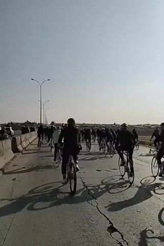 A group of people ride bicycles on a paved road, forming a pack of cyclists moving together. The road appears to be a bridge or overpass, with a concrete barrier on the left side, separating the bike lane from traffic. Several cars are visible on the other side of the barrier. Streetlights line the road. The road surface shows cracks and signs of wear. The sky is clear and bright, indicating a sunny day, and the cyclists are casting long shadows on the road. Most of the riders are wearing dark clothing.