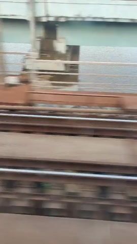 The video shows a view from a moving train looking out a window. In the foreground are train tracks. In the middle ground, there is a bridge with railings and support beams. The bridge crosses over a body of water. In the background, there are green hills, some vegetation, power lines, and a cloudy sky. The overall impression is that of a scenic train journey.