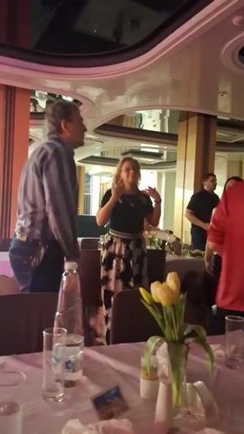 Several people are dancing in what appears to be a restaurant or event space. There are tables set with white linens, glasses, water bottles, and small bouquets of yellow tulips. The man in the foreground is wearing a blue striped shirt and jeans. A woman in a black top and black-and-white patterned skirt is dancing beside him. Other people are dancing in the background. The space has a mirrored ceiling and pillars. The lighting changes from neutral to blue.