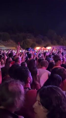 The video captures a crowd of people at an outdoor event at night, illuminated by purple and pink stage lighting. Many individuals are holding up their cell phones, likely recording or taking pictures, with some people raising their hands in the air. The audience appears to be densely packed together, facing towards a stage or performance area that is not fully visible. The background consists of a dark sky and a sloping hillside with some vegetation. The overall atmosphere suggests a lively concert or festival.