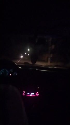 The video shows the interior of a car being driven at night. The dashboard and radio are illuminated, and the driver's arm is visible. The car is moving forward on a road with streetlights and other vehicles in the distance. A traffic sign is visible on the right side of the road. The overall scene is dark, with the primary illumination coming from the car's dashboard and the headlights of other vehicles.