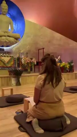 The video shows the interior of what appears to be a Buddhist meditation hall. A woman sits on a cushion on the wooden floor, facing forward. She has medium-length brown hair styled in a low ponytail, and she is wearing a beige shirt and patterned skirt. In front of her is a small wooden stand with papers on it. To her left and right are other meditation cushions spaced apart. In the background, a raised platform holds an altar decorated with flowers, candles, and other religious items. A statue of Buddha is prominently displayed on the left, illuminated with blue light. A person in traditional robes is seated at the front of the platform, appearing to lead the meditation. The walls are decorated with earth-toned textured surfaces. The scene suggests a moment of quiet contemplation and guided meditation in a spiritual setting.