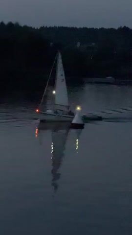 A sailboat is moving through dark water at dusk. The boat has a white sail with a number "4" visible on it. The boat is equipped with running lights. There is a person on the boat. A marker buoy is visible in the water next to the boat. The water is calm and reflects the lights from the boat. The background shows a dark treeline with some lights from houses or buildings. The overall scene is peaceful and serene.