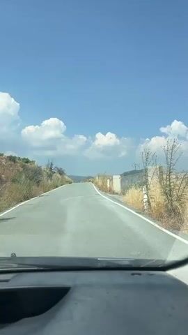 The video shows a view from inside a car looking out through the windshield at a two-lane asphalt road stretching into the distance under a clear blue sky with scattered white clouds. The road has a white line on the right edge. The landscape on either side of the road is dry and sparse, with brown grass and some green bushes on the left, and brown grass, some bushes, and a concrete structure with a wire fence on the right. The road appears to be slightly uphill and curves gently to the right in the distance. The bottom of the frame shows the car's dashboard. The timestamp indicates the video is a short clip, showing minimal movement of the car.