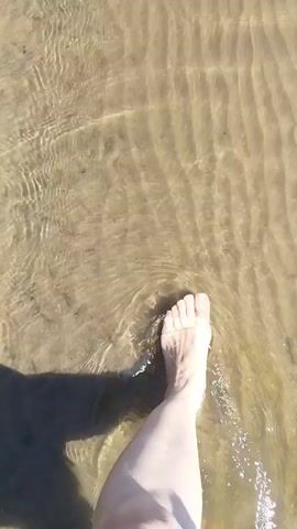 The video shows a person's bare foot walking through shallow, clear water over a sandy, rippled seabed. The camera angle is from above, looking down at the foot. The water is casting ripples and reflections on the sand.