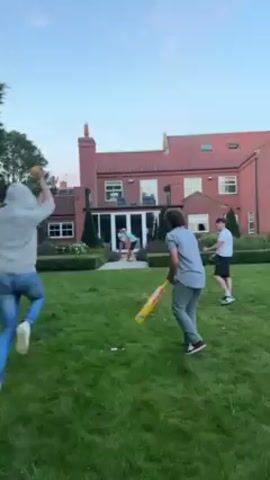 The video shows four young men playing cricket on a lawn in front of a large brick house. The house has a red roof and several windows. The lawn is green and well-maintained, with hedges and a few trees. One man is throwing a cricket ball, and another man is holding a yellow plastic cricket bat. The other two men are standing nearby. The man with the bat trips and falls. The man who threw the ball runs toward the fallen man. It is evening, and the sky is clear.