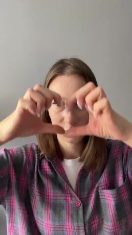 A woman with shoulder-length brown hair and fair skin is centered in the frame, looking directly at the camera. She is wearing a pink and gray plaid button-up shirt over a white t-shirt. Her hands are brought up to her face, forming a heart shape around her eyes. The background is a plain, light gray wall. The lighting is even and comes from the front. The woman's facial expression is neutral to slightly smiling. Her fingernails are painted white.