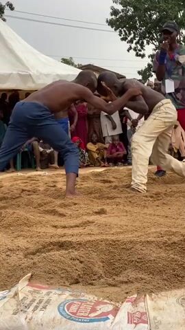 Two young, shirtless boys wrestle on a sand-covered surface. They are surrounded by a crowd of onlookers of varied ages and dress. There is a white tent in the background. One of the boys is wearing blue pants, the other light tan. A man in a black hat and camouflage shirt oversees the wrestling match. The boys grapple, maneuvering around each other. Bags of plaster line the edge of the sand pit. Trees are visible in the background. The overall environment suggests an outdoor event or competition.