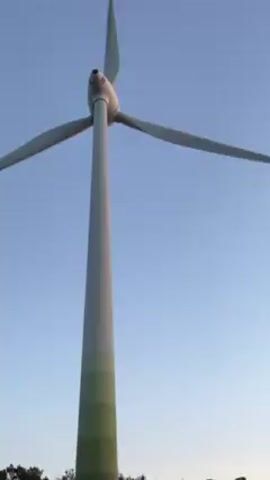 The video shows a wind turbine spinning against a clear blue sky. As the camera zooms out, the base of the turbine is revealed, painted with green stripes and some graffiti. A vintage-style green and white van is parked in the background.  Trees are visible along the horizon.