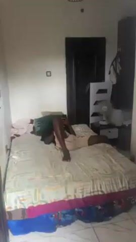 A person lies face down on a bed in a small bedroom, using a phone. The room has white walls, a tiled floor, and is sparsely furnished. A dark wood door is visible, along with a wardrobe, a bedside table with a fan, and a few other items. The bed has patterned sheets and a green pillow. The person is wearing light-colored shorts.