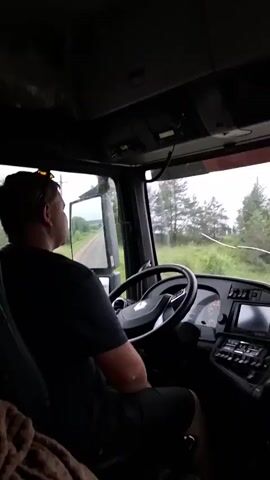 A person sits in the driver's seat of a truck, facing forward. The person is wearing a black t-shirt and dark shorts. The truck's interior is visible, including the steering wheel, dashboard, and various controls. Outside the windshield, a road can be seen stretching into the distance, bordered by trees and greenery. The sky appears overcast. The video is shot from inside the truck, looking towards the driver and the road ahead.