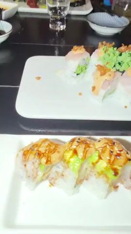 The video shows two plates of sushi on a table. The top plate has several pieces of sushi rolls topped with sauce and some green garnish. The bottom plate has more sushi rolls, which appear to be unagi rolls, covered in sauce and topped with sesame seeds and avocado. There are also small dishes with pickled ginger and wasabi on the side. In the background, there are drinks and other food items on the table, and a dark, possibly restaurant setting. The camera is focused on the sushi dishes, and the lighting is dim.