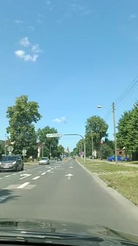 The video shows a forward-facing view from a vehicle driving down a paved road lined with trees and houses. The sky is blue with a few white clouds, indicating it is daytime and the weather is clear. The road has multiple lanes and white lane markings. A speed limit sign shows "40". A signpost overhead indicates "Centrum Wodne LAGUNA" with an arrow pointing to the left, suggesting a water park or aquatic center. Other traffic signs are visible, including a "no stopping" sign. Houses with red-tiled roofs are visible on the left side of the road, and more trees and houses are on the right. The scene appears to be in a suburban or town setting. The overall atmosphere is calm and ordinary, as the vehicle moves through the town.