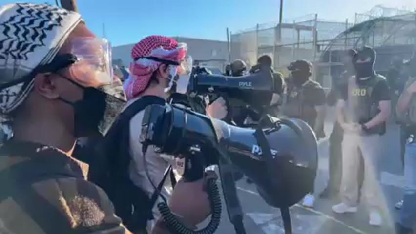 The video shows an outdoor protest or demonstration in Newark, United States, likely outside a detention center or secure facility, indicated by a chain-link fence topped with barbed wire visible in the background. The scene is brightly lit by the sun.

In the foreground, two individuals are prominent among a group of protesters, each holding a black megaphone branded "Pyle." One protester wears a Keffiyeh, clear safety goggles, and a black face mask, actively chanting into the megaphone. Another protester next to them also holds a megaphone and wears a red Keffiyeh. They are heard chanting in Spanish, including the phrase "es pura porquería" (it's pure filth).

Facing the protesters is a line of approximately ten individuals dressed in tactical gear. All of them wear black balaclavas that obscure their faces, tactical vests, and various pants (jeans, khaki, camouflage). Several of the vests clearly display the letters "ERO," while at least one vest has "POLICE ICE" written on it. They stand stoically and observe the protesters. Behind them and to the right, a large, dark green armored vehicle, resembling an MRAP, is parked. The overall atmosphere appears tense, with vocal protesters engaging silent, armed personnel.