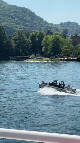 A wide, long shot depicts a boat moving quickly across a lake on a sunny day. Several passengers are aboard the boat, which is leaving a long wake behind it.  In the background, there's a lush green hill and buildings on the shore.  A white railing is visible in the foreground, suggesting the perspective is from another boat.  The sky is clear and blue.