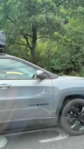 A gray Jeep Compass with a roof tent is driving on a paved road next to a wooded area. The Jeep is towing a trailer with a white and blue BMW motorcycle secured to it. The motorcycle has red accents on the seat and blue straps holding it in place. The video pans from the front of the Jeep to the back, showing the motorcycle in detail.