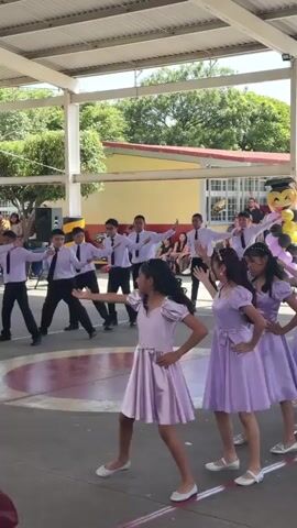 A group of young people, likely students, are performing a coordinated dance routine outdoors. The performers are divided into two apparent groups: one group consists of boys in light purple collared shirts and black ties with black pants, and the other group consists of girls in matching light purple dresses. The girls' dresses are knee-length with short, puffed sleeves and sashes tied around their waists. They are all wearing white ballet flats. The boys' attire is consistent across the group, and they all appear to be wearing black dress shoes. The performance is taking place on a paved surface, possibly a school courtyard or gymnasium, under a covered roof structure. In the background, a building with yellow and red accents is visible, along with trees and an audience of spectators, some of whom are holding up phones to record the event. The lighting suggests it is daytime, with sunlight filtering through the roof. There are speakers visible on the left side of the frame. One individual in the foreground, in the lower left corner, is holding a phone, suggesting they are filming the performance. The overall atmosphere appears to be one of a school event or performance.