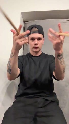 A young man with tattoos on his forearms sits and spins drumsticks in his hands. He wears a black t-shirt, black pants, and a black baseball cap worn backwards. He has fair skin and short blond hair. His arms are raised and his hands are extended, palms facing forward, as he twirls the drumsticks between his fingers. He appears focused and is looking directly at the camera. He has headphones in his ears. The background consists of a white wall on the left and a mottled gray surface on the right.
