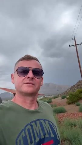 This is a selfie video of a man wearing sunglasses and a green t-shirt. He is outdoors with a cloudy sky. In the background, there are mountains, vegetation, a utility pole with wires, and a rest area with a picnic table. The man smiles and gives a thumbs up.