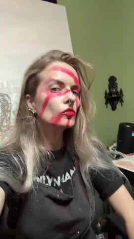 A woman with long blonde hair, fair skin, and blue eyes is looking directly at the camera. She has bright red makeup smeared across her face in thick, uneven stripes and has a red lipstick applied. She has several piercings on her left ear and a septum piercing. She is wearing a black T-shirt with white lettering that is partially obscured by a black apron. The background consists of a green wall with a framed religious icon, a partially visible canvas with a painting in progress, a black decorative wall sconce, and various painting supplies. The lighting appears to be artificial and somewhat dim.