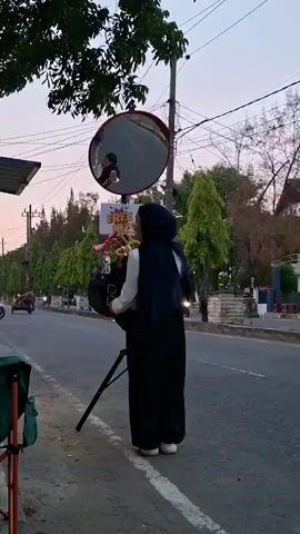 A woman in a hijab and a white blouse with black pants stands on the side of a road, holding a bouquet of flowers. She is facing a large, circular convex mirror mounted on a pole. In the reflection of the mirror, the woman can be seen holding her phone to her face, seemingly taking a selfie. A sign with the words "FREE PHOTO" is visible behind the bouquet and below the mirror. The scene takes place outdoors with trees lining the road and buildings in the background. The sky appears to be dusky. A tripod is set up near the woman, supporting the bouquet and possibly a camera.