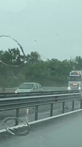 The video captures a view through a wet windshield from a moving vehicle on a highway during overcast weather. In the foreground, raindrops are visible on the glass, distorting the view. A metal guardrail runs along the side of the highway. In the middle ground, a silver Volvo semi-truck with a trailer is traveling on the highway. The truck has lights on and an amber warning light flashing on its roof. Further in the distance, a billboard for McDonald's is visible, displaying the iconic golden arches logo and the text "80 km" with an upward-pointing arrow. The background consists of a dense line of trees. The overall impression is of a highway journey on a rainy day.