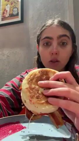 A young woman with brown hair braided into pigtails eats a sandwich. She is wearing a red and grey striped shirt. The sandwich has a round, toasted bun, and bacon and other ingredients are visible inside. She takes a large bite, looking directly at the camera. A blue and red patterned plate sits beneath the sandwich. A framed picture hangs on the grey wall behind her. The picture features a cat and a clock. There are mechanical sounds audible in the background.