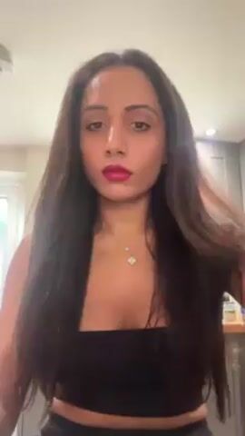 A woman with long brown hair, dark eyes, and wearing dark pink lipstick is standing indoors. She has a nose ring and is wearing a black top and a necklace. She is drinking from a white mug, then she smiles at the camera. A person can be seen briefly in the background.