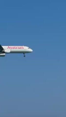 Here is a description of the video:

An Austrian Airlines passenger plane is descending towards the ocean. It appears to be landing at an airport close to a beach, as people are standing in the water in the foreground. The sky is blue and clear.