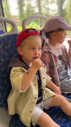 Two young children are sitting on a blue bus seat, looking out the window. The child on the left is a toddler wearing a red baseball cap, a khaki jacket, a dark shirt, and gray shorts. They have a lollipop in their mouth and a small stain on their arm. The child on the right is slightly older, wearing a light purple baseball cap, a pink plaid shirt over a white t-shirt, and denim shorts. They also have a lollipop in their mouth. Both children are holding hands. The background shows trees and buildings passing by outside the bus window. The bus seat has a blue speckled pattern.